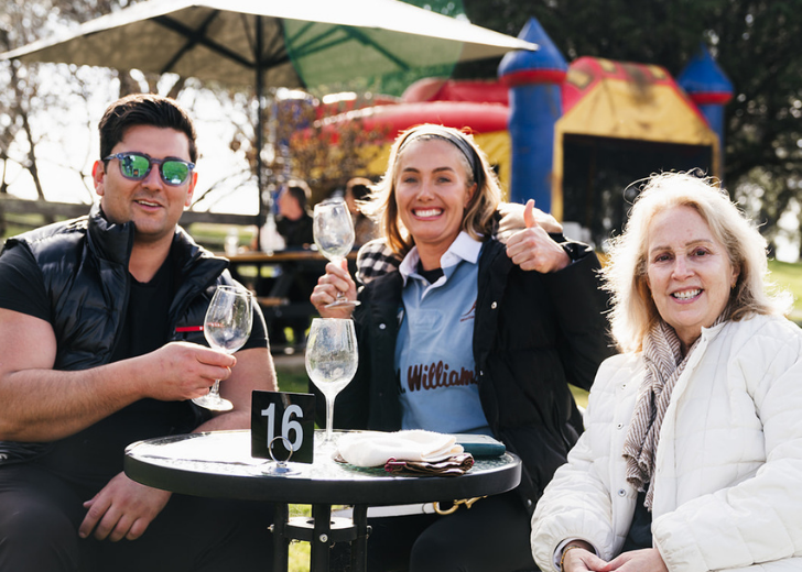 Three people sitting at a table outdoors with wine glasses, in front of a playground.