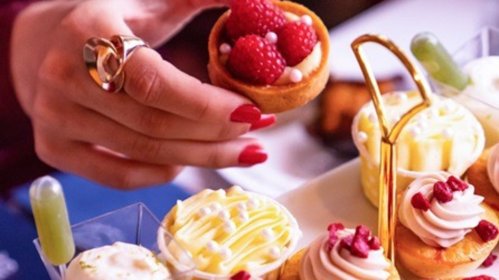 Hand with red nail polish holding a tartlet with raspberries over a display of pastries.