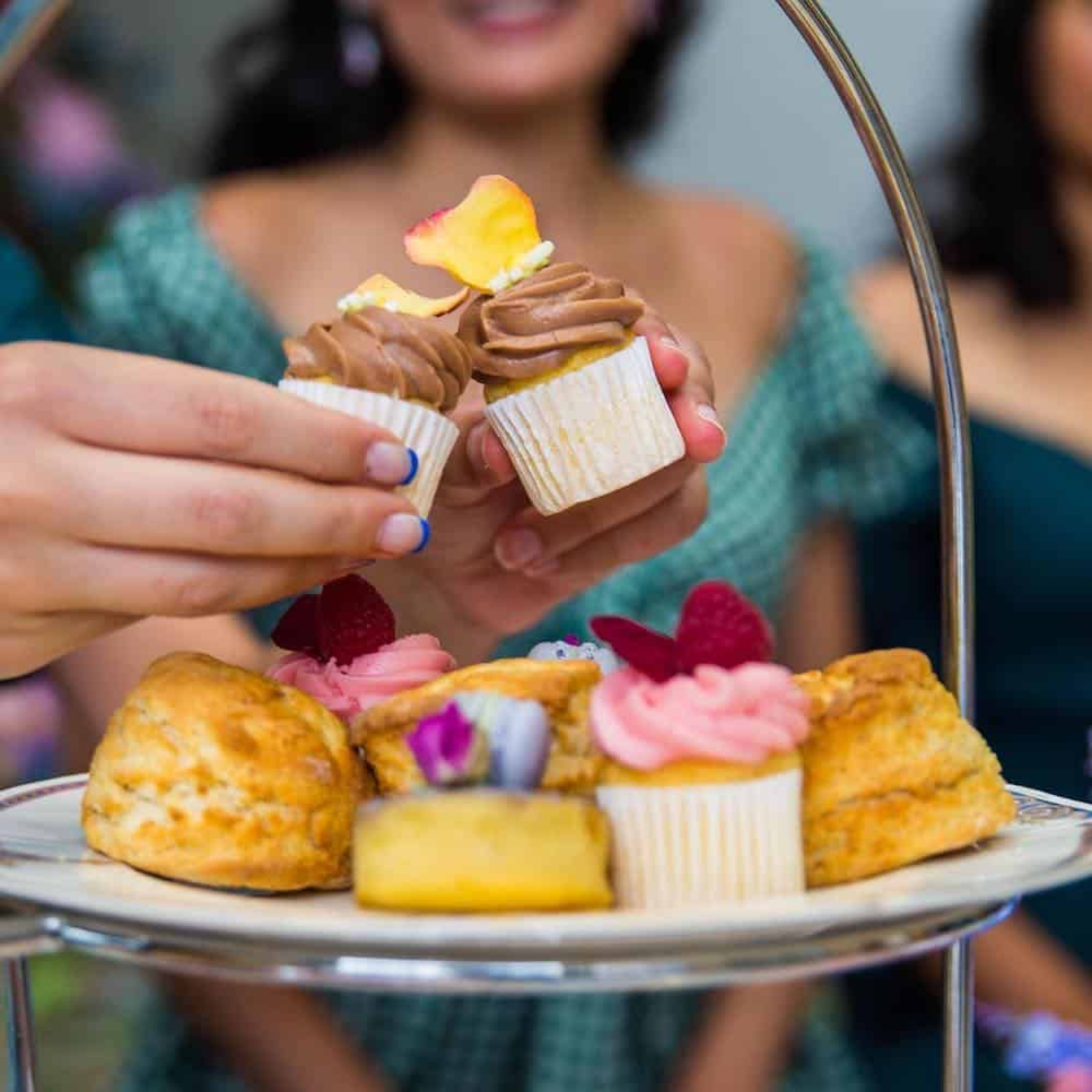 Person holding a cupcake with high tea above a tiered cake stand with pastries and more cupcakes.