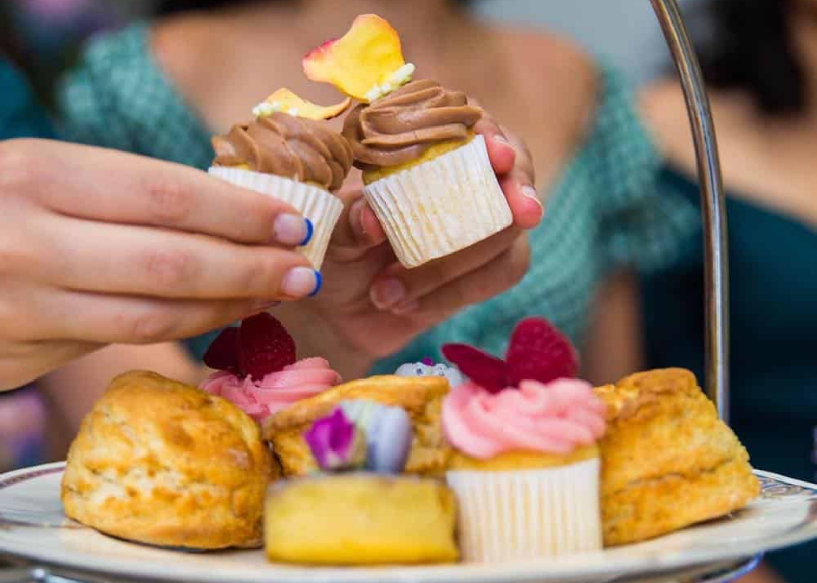 Person holding a cupcake with high tea above a tiered cake stand with pastries and more cupcakes.