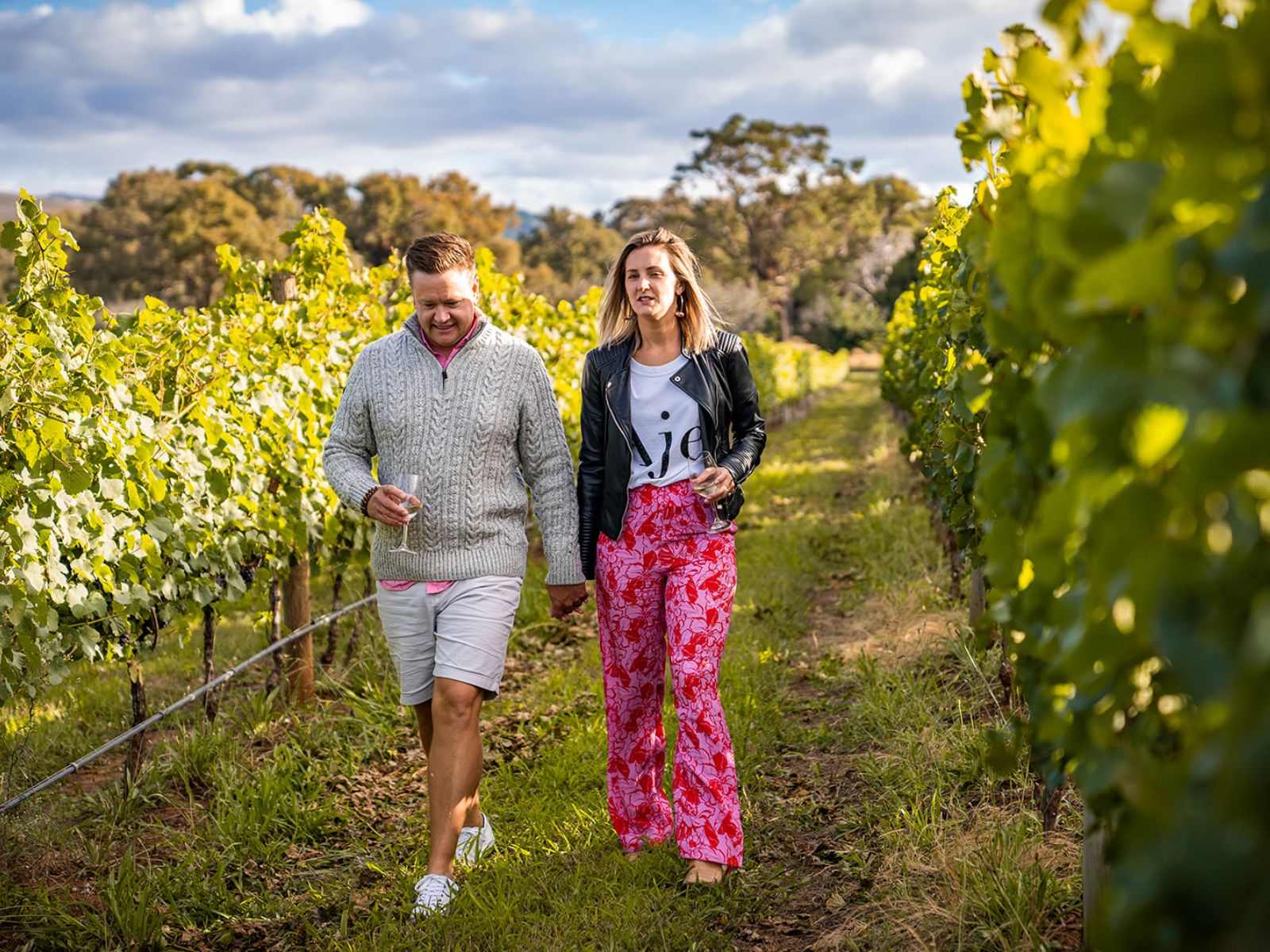 A male and female couple holding hands and glasses of wine while walking through a vineyard at Stockman's Ridge Wines