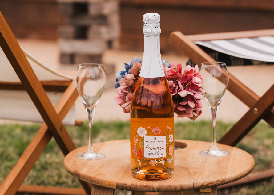 A bottle of sparkling rosé wine with a white foil top and a floral orange label sits on a small wooden table outdoors, flanked by two champagne flutes and pink hydrangeas. Lounge chairs and a wooden deck are blurred in the background.