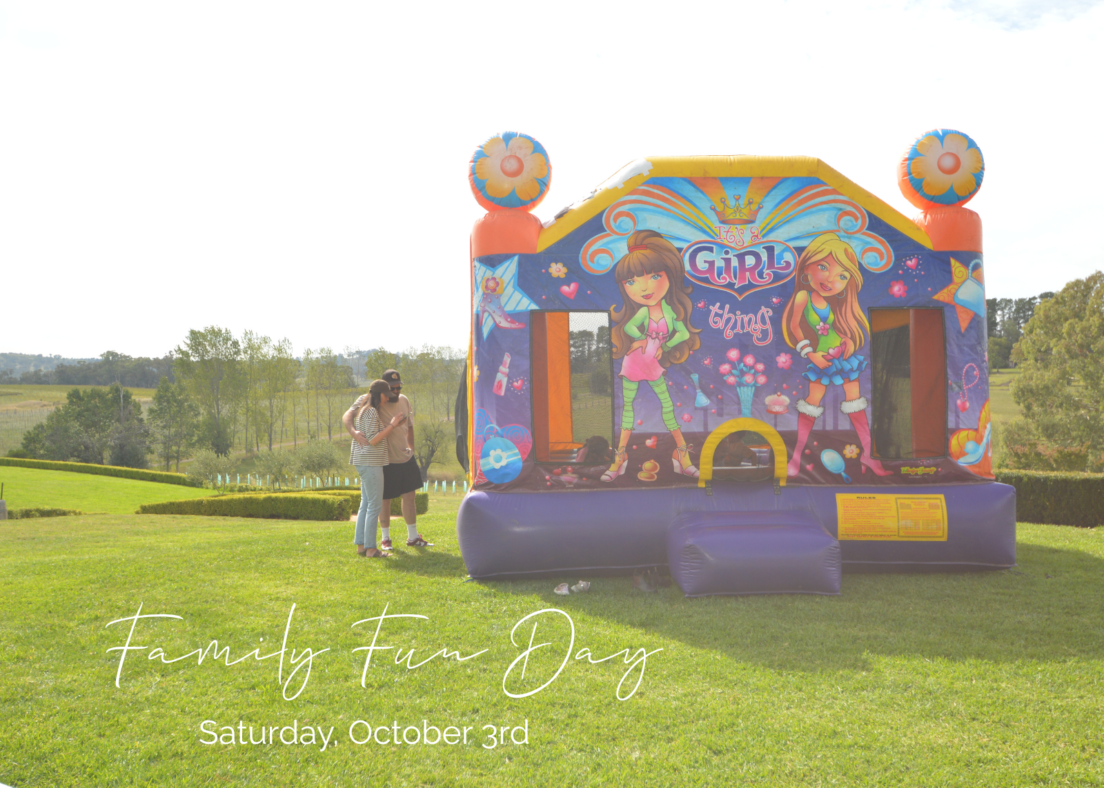 A colorful inflatable children's bounce house with cartoon girl graphics stands on a grassy lawn outdoors. Two people are standing near the left side of the bounce house, and the sky is bright and overexposed.