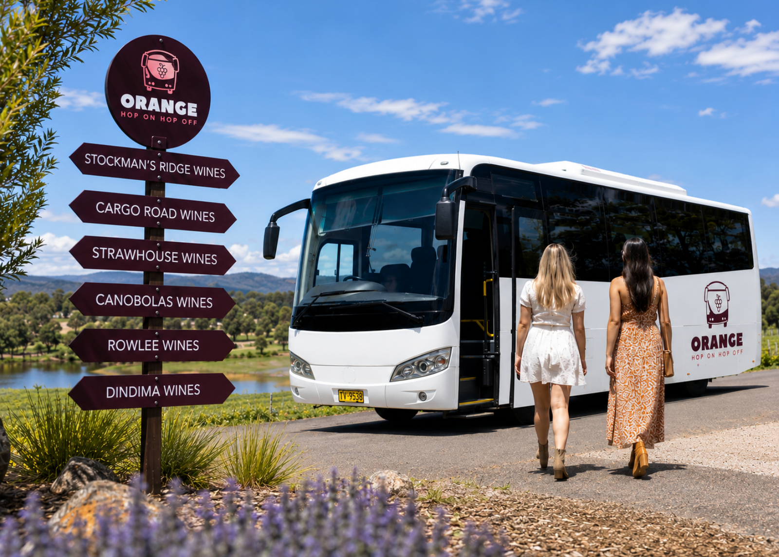 A white Orange Winery tour bus parked outdoors beside a signpost listing wine estates, with two women walking toward the open passenger door. The scene includes a blue sky, water, and landscaped roadside setting.