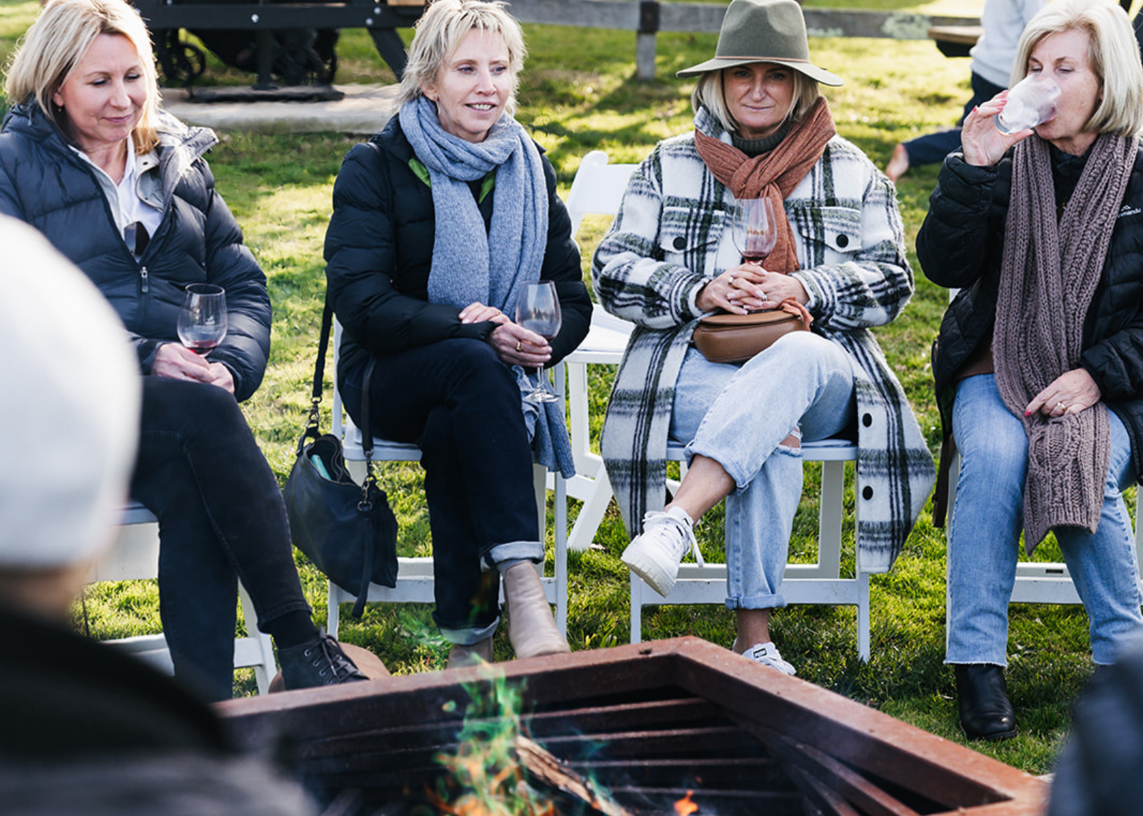 Four women sitting around a fire pit in a park-like setting, enjoying drinks.