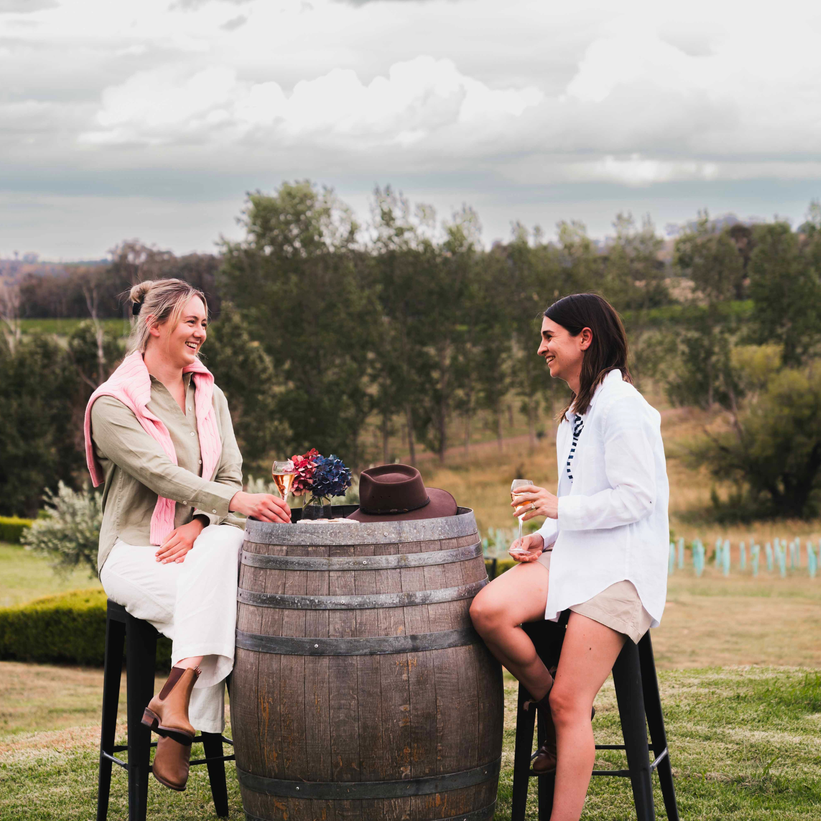 Two women sitting on stools around a barrel in an outdoor setting with trees and open space in the background at Stockman's Ridge Wines