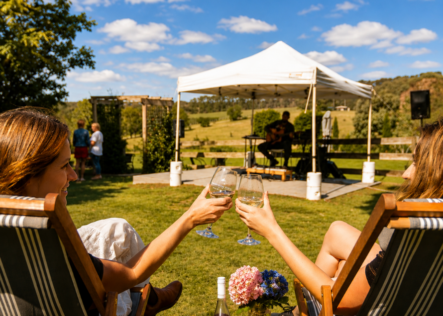 Two women sitting in striped deck chairs clinking wine glasses outdoors at a sunny vineyard or garden event, with a white canopy, musicians, grass, and blue sky in the background.